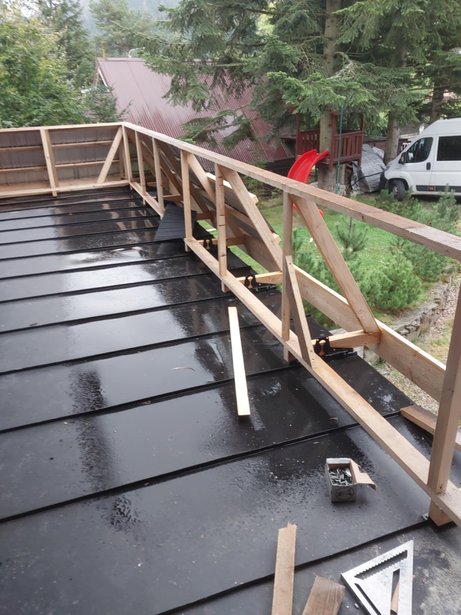 Czarny, mokry dach z blachy trapezowej w trakcie montażu drewnianej konstrukcji, widoczne narzędzia i pudełko ze śrubami, w tle drzewa i zabudowania.