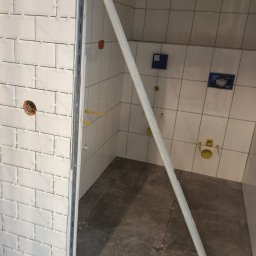 Zbigniew Dziura - Łazienka w trakcie wykańczania: białe płytki ścienne z krzyżykami dystansowymi, szare płytki podłogowe, widoczne instalacje wodno-kanalizacyjne i otwór elektryczny, zabezpieczona listwą krawędź...