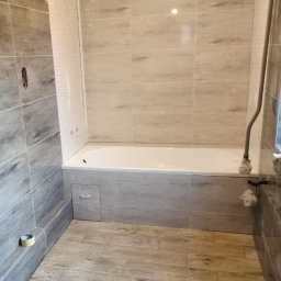 Zbigniew Dziura - Wykończona łazienka w Krosno z wanną obudowaną płytkami imitującymi drewno, pasującymi do podłogi, jasne płytki na ścianach, widoczna rolka taśmy malarskiej przy podłodze.