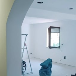 Zbigniew Dziura - Wnętrze w trakcie malowania: widoczna drabina, zakryty worek z odpadami, częściowo pomalowane ściany na biało, sufit z zamontowanymi punktami świetlnymi, otwarte okno z roletami.