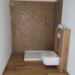 Zbigniew Dziura - Łazienka w trakcie remontu: mozaika w odcieniach brązu na ścianie prysznicowej, biały brodzik, podwieszana toaleta zabezpieczona folią, drewnopodobna podłoga, sufit z zamontowanymi punktami...