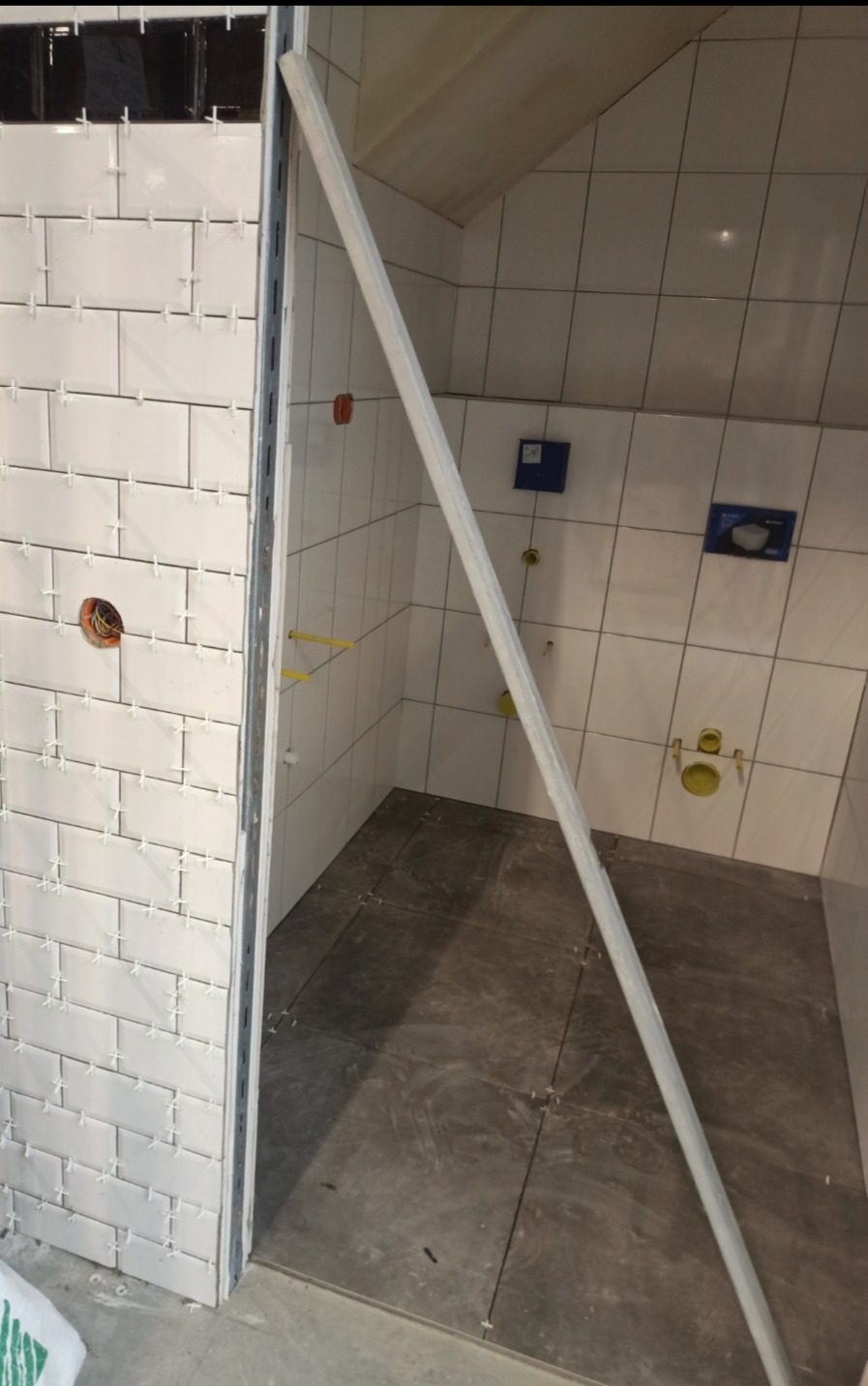 Łazienka w trakcie wykańczania: białe płytki ścienne z krzyżykami dystansowymi, szare płytki podłogowe, widoczne instalacje wodno-kanalizacyjne i otwór elektryczny, zabezpieczona listwą krawędź...