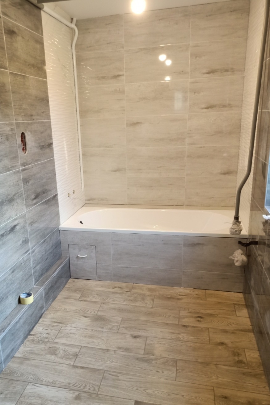 Wykończona łazienka w Krosno z wanną obudowaną płytkami imitującymi drewno, pasującymi do podłogi, jasne płytki na ścianach, widoczna rolka taśmy malarskiej przy podłodze.