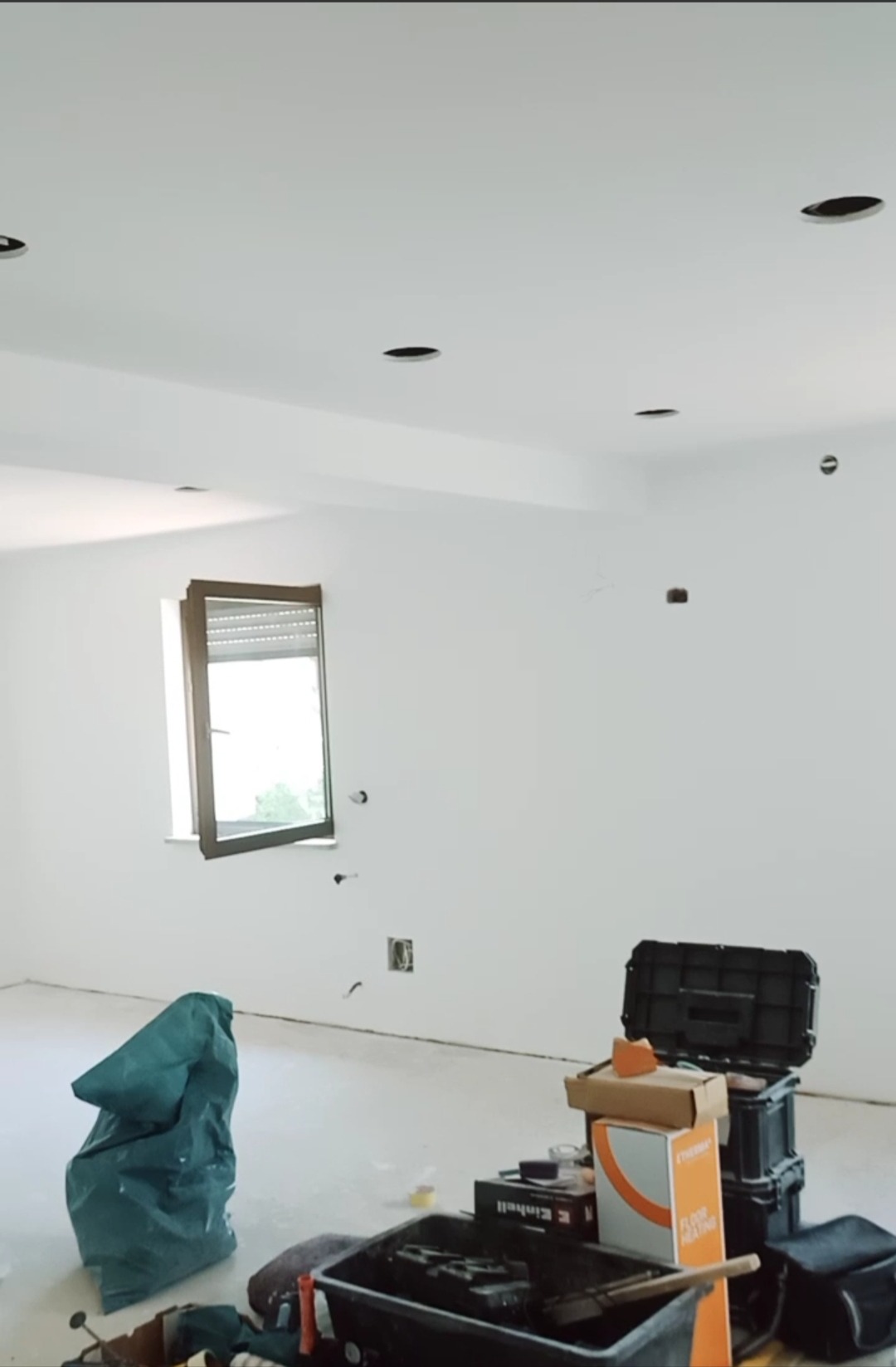 Wnętrze pokoju w trakcie remontu: białe ściany z widocznymi otworami na oświetlenie, okno z roletą, narzędzia i materiały budowlane na podłodze, w tym wiadro, skrzynka narzędziowa i opakowanie po...