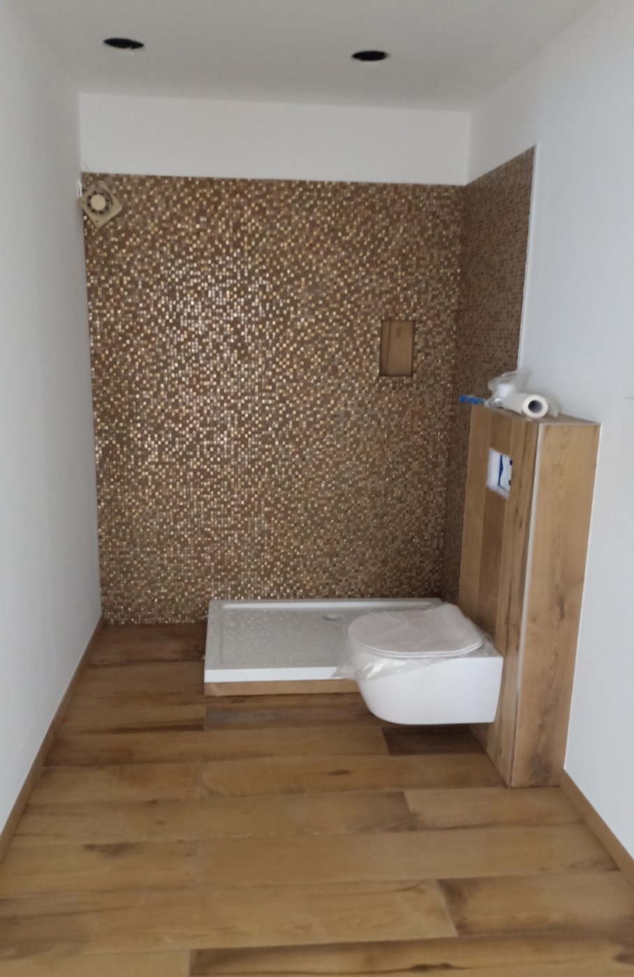 Łazienka w trakcie remontu: mozaika w odcieniach brązu na ścianie prysznicowej, biały brodzik, podwieszana toaleta zabezpieczona folią, drewnopodobna podłoga, sufit z zamontowanymi punktami...