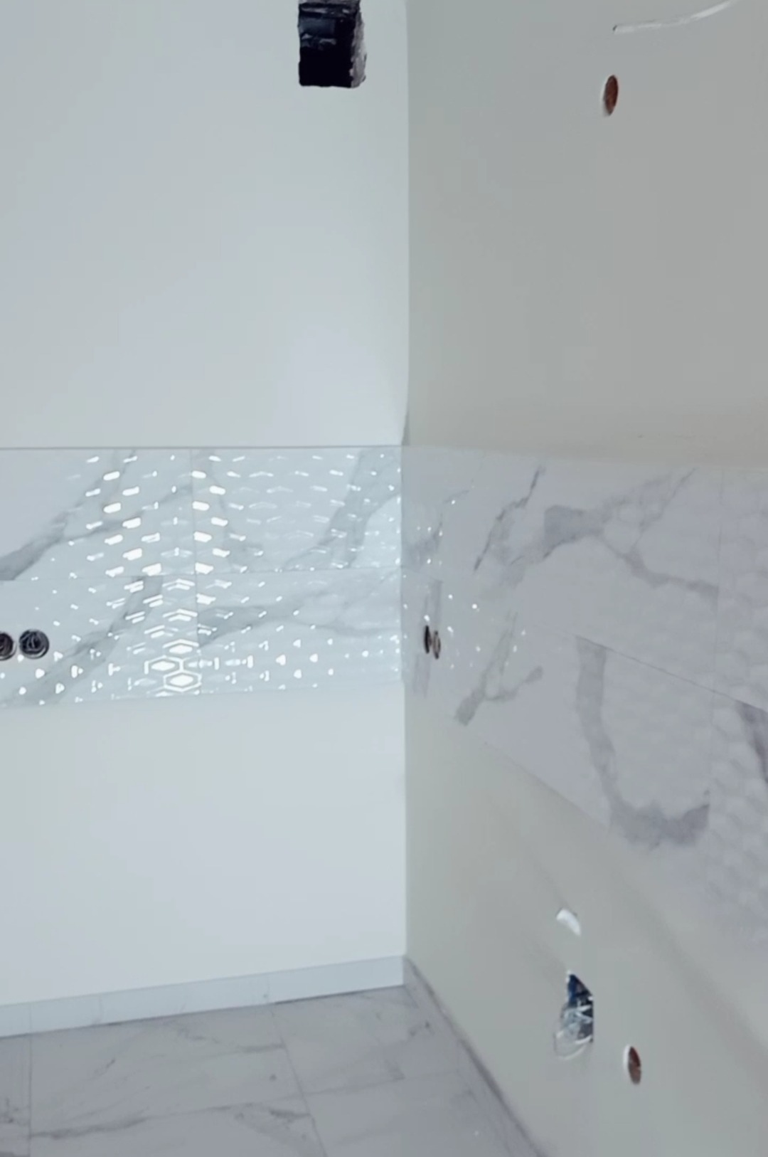 Ściana w trakcie wykańczania z częściowo ułożoną biało-szarą glazurą o wzorze marmuru i heksagonów, widoczne otwory na gniazdka elektryczne i puszki instalacyjne.