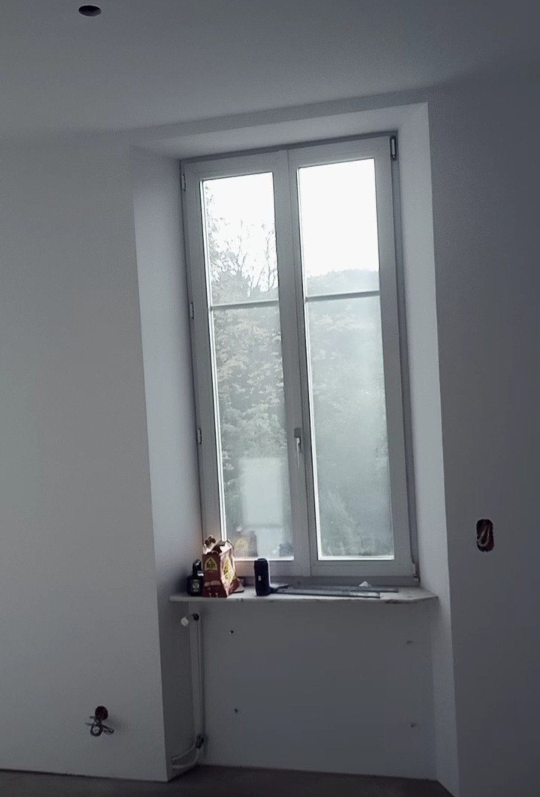 Wnętrze pokoju po świeżym pomalowaniu na biało, widoczne okno z widokiem na drzewa, parapet z akcesoriami, grzejnik i gniazdko elektryczne bez osłonki.