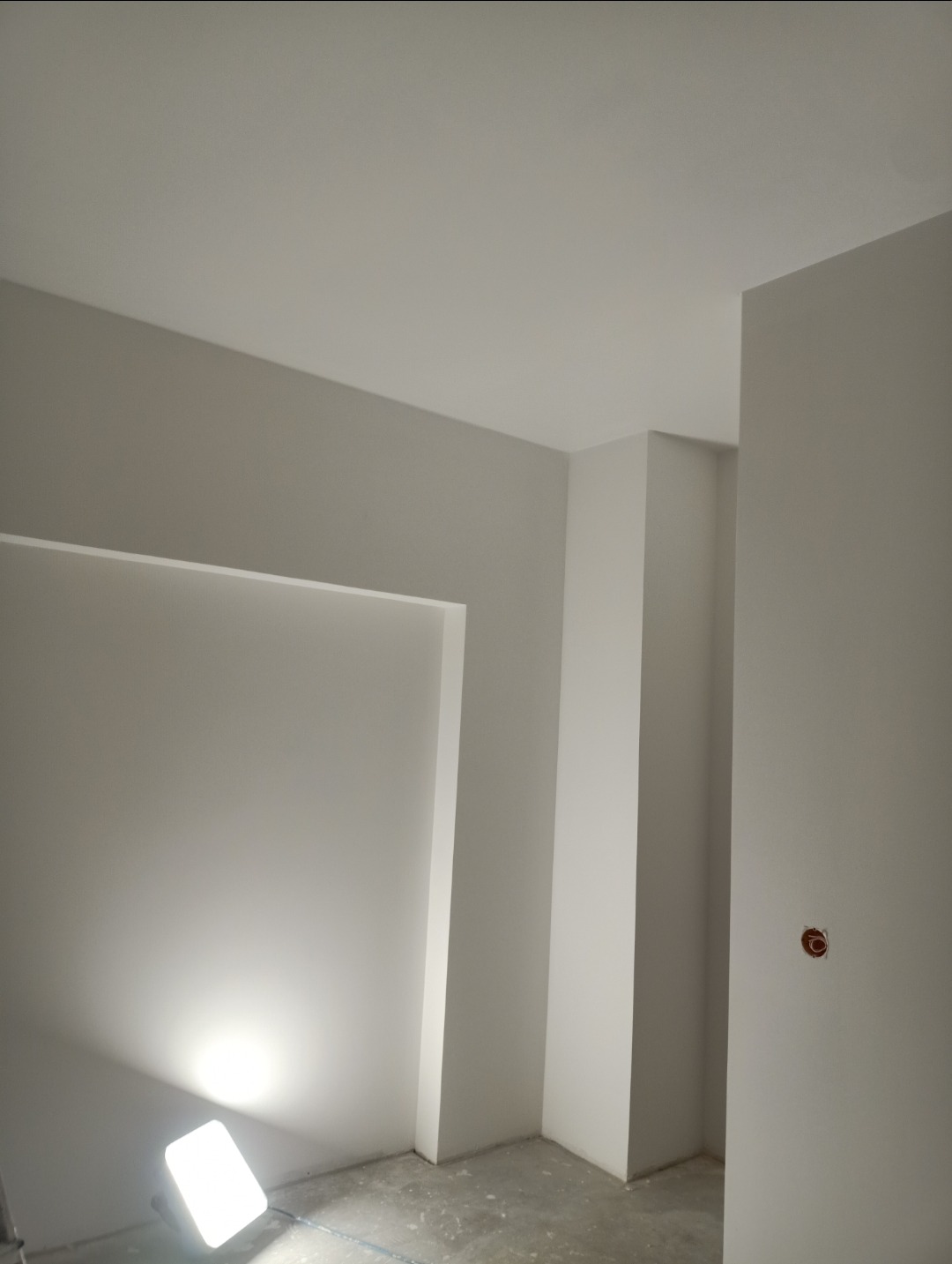 Wnętrze w trakcie remontu, widoczne ściany przygotowane do malowania lub wykończenia gładzią, z lampą halogenową oświetlającą fragment ściany.