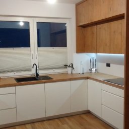 Marcin Konkol - Nowoczesna kuchnia w odcieniach bieli i drewna z czarnym zlewem i baterią, oświetleniem podszafkowym LED i roletami okiennymi.