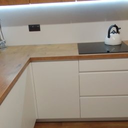 Marcin Konkol - Jasna kuchnia z drewnianym blatem, białymi frontami szafek i czarną płytą indukcyjną, uzupełniona o biały czajnik i akcesoria kuchenne.