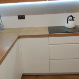 Jasna kuchnia z drewnianym blatem, białymi frontami szafek i czarną płytą indukcyjną, uzupełniona o biały czajnik i akcesoria kuchenne.