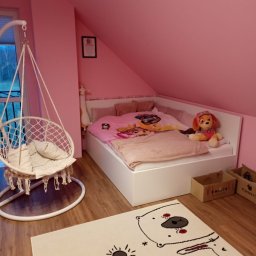 Marcin Konkol - Pokój dziecięcy na poddaszu z różowymi ścianami, białym łóżkiem w zabudowie, wiszącym fotelem hamakowym i dywanem z motywem polarnym.