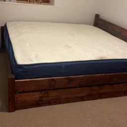 Łóżko z ciemnego drewna z materacem w pokoju z oknem, na parapecie dekoracja 'Relax'