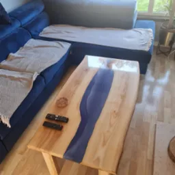 Stolik kawowy z blatem z jasnego drewna z żywicą epoksydową w kolorze granatowym, obok sofy z poduszkami i pilotami.