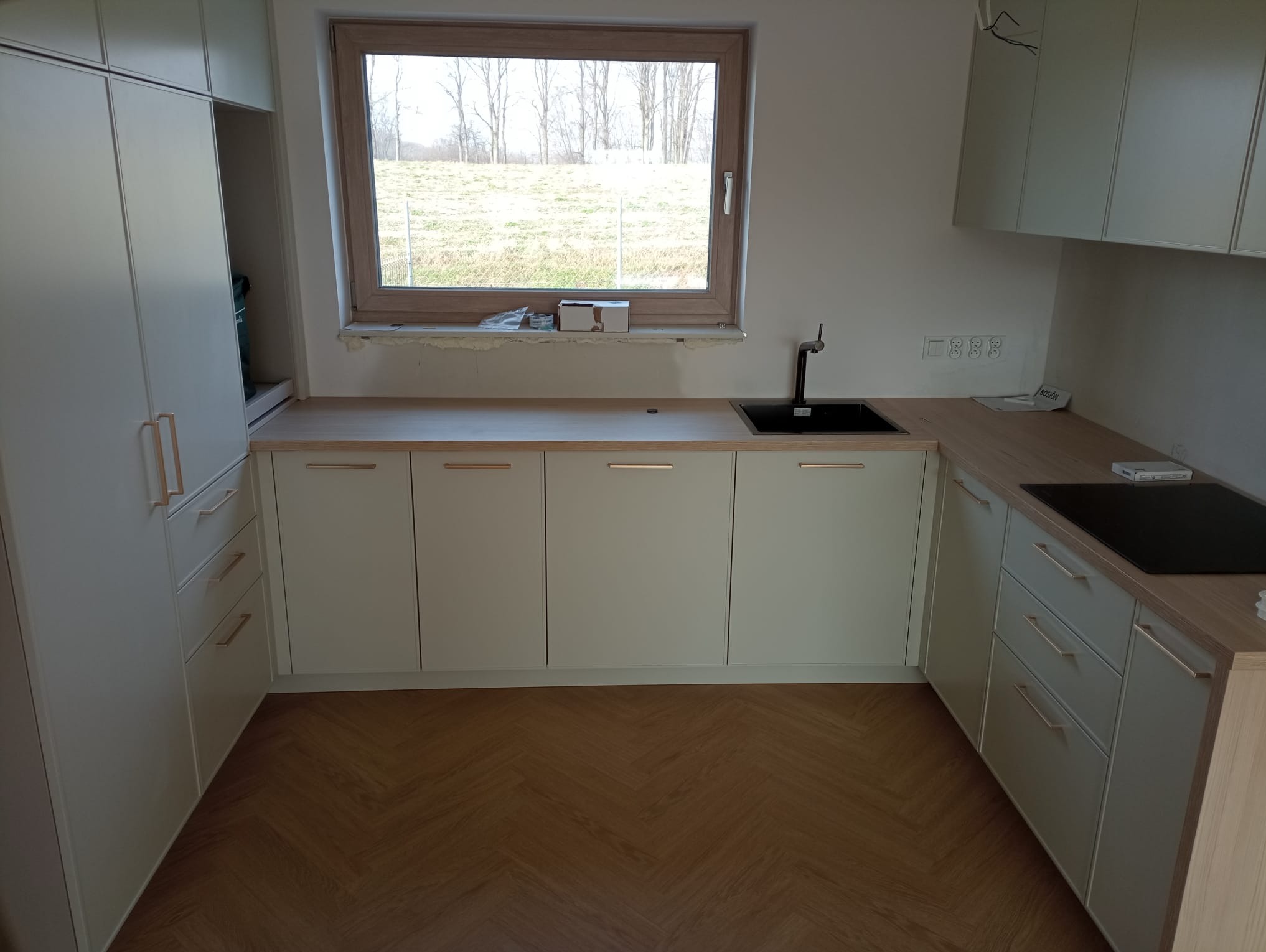 Nowoczesna kuchnia z jasnymi frontami szafek i drewnianym blatem, czarny zlew wpuszczany, widok na krajobraz za oknem, parkiet ułożony w jodełkę.