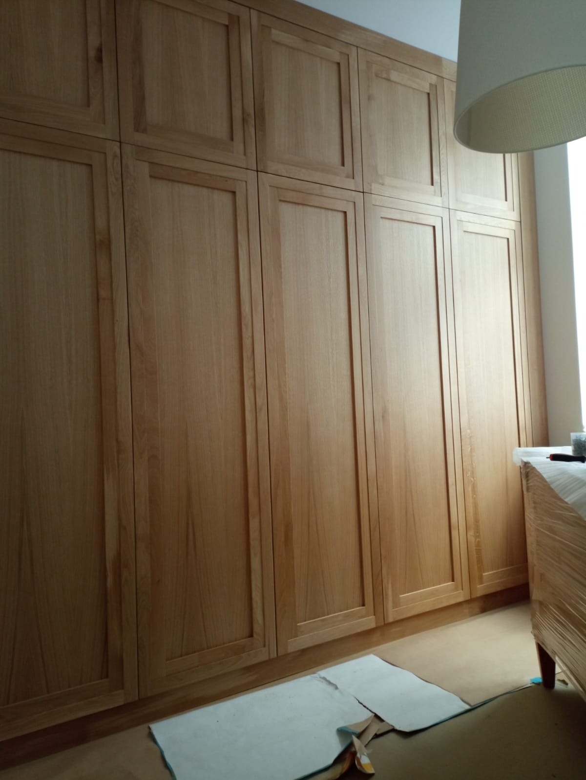 Wysoka, drewniana szafa wnękowa z jasnego drewna, z widocznymi ramkami na frontach, częściowo zakryta przez lampę i mebel zabezpieczony folią.