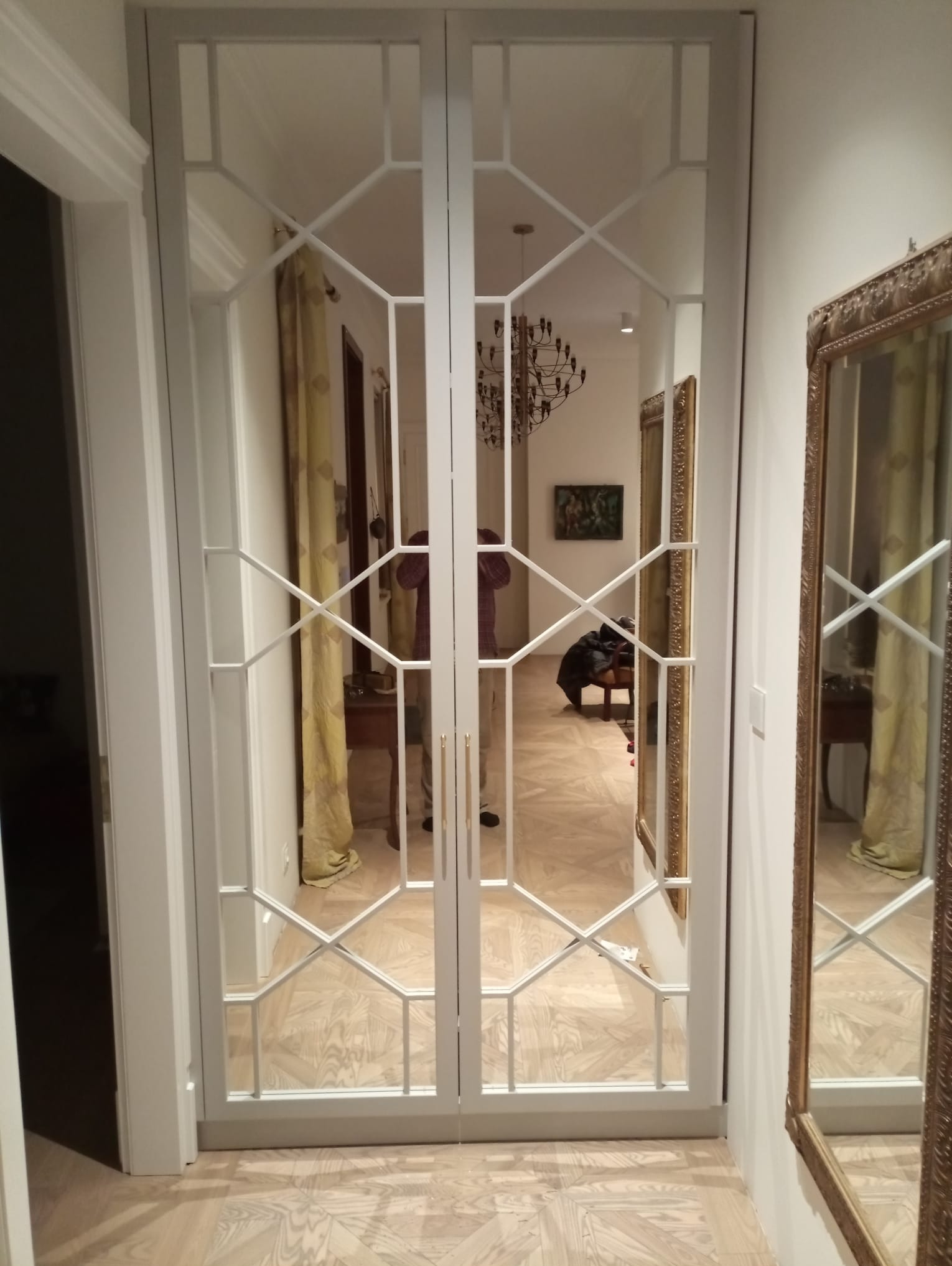 Białe, przeszklone drzwi w geometryczne wzory, odbijające wnętrze z parkietem i żyrandolem, widoczne lustro z ozdobną ramą po prawej stronie.