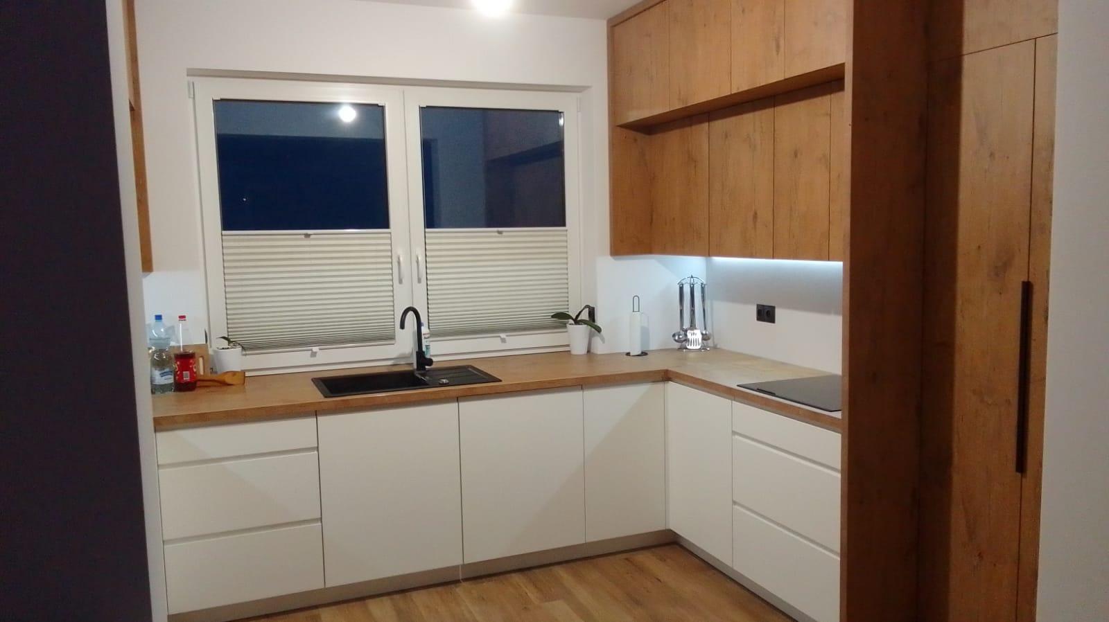 Nowoczesna kuchnia w odcieniach bieli i drewna z czarnym zlewem i baterią, oświetleniem podszafkowym LED i roletami okiennymi.