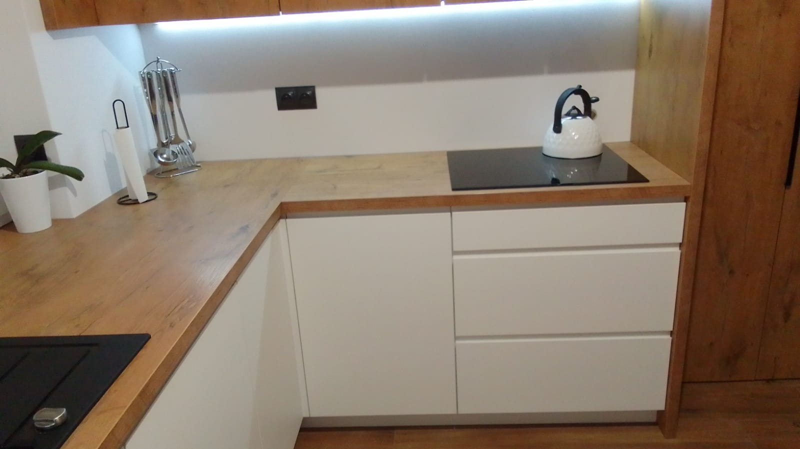 Jasna kuchnia z drewnianym blatem, białymi frontami szafek i czarną płytą indukcyjną, uzupełniona o biały czajnik i akcesoria kuchenne.