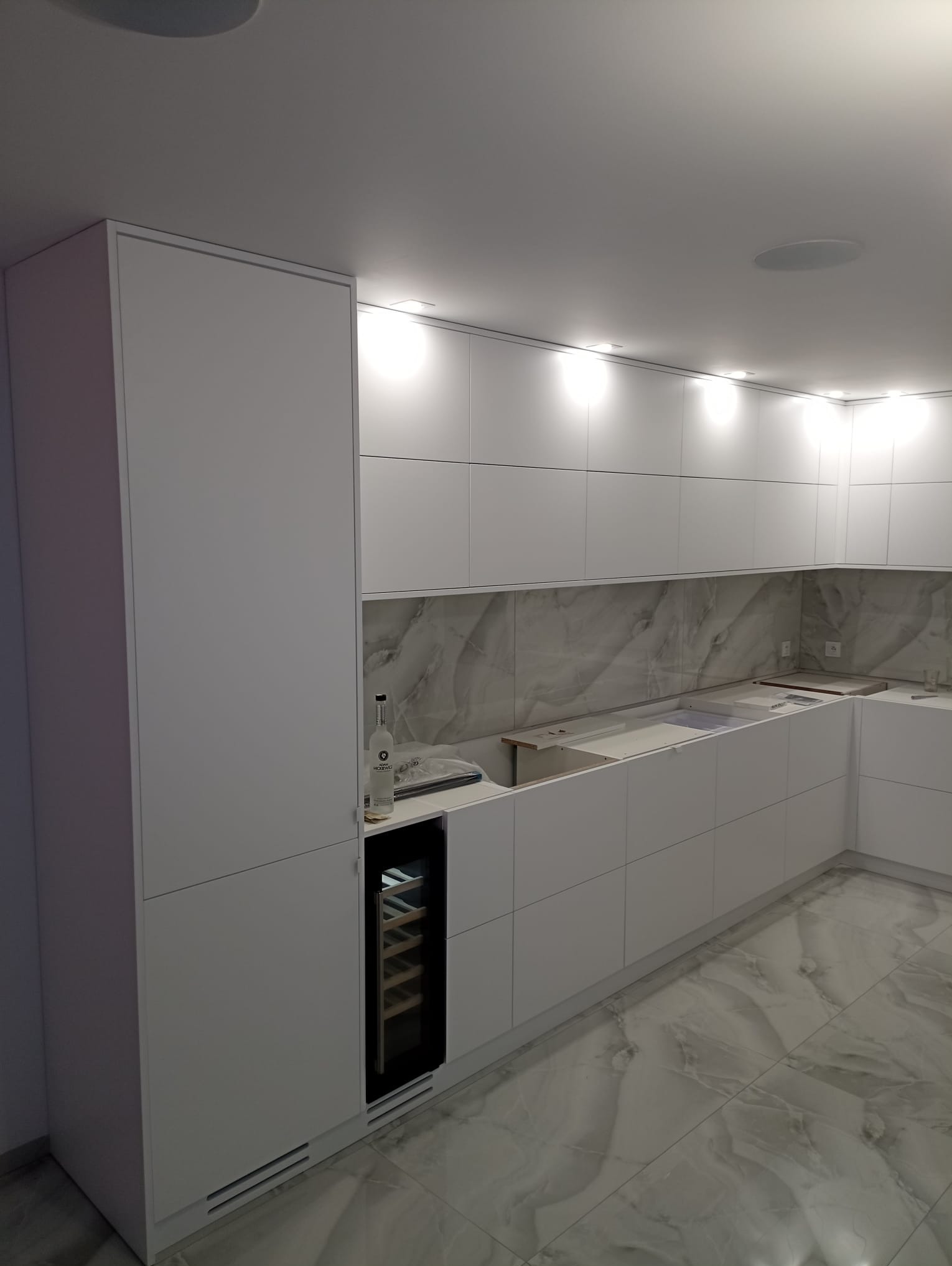 Nowoczesna, biała kuchnia na wymiar w trakcie montażu, z marmurowym backsplashiem i podświetleniem LED, wyposażona w wbudowaną chłodziarkę do wina i butelkę wódki na blacie.