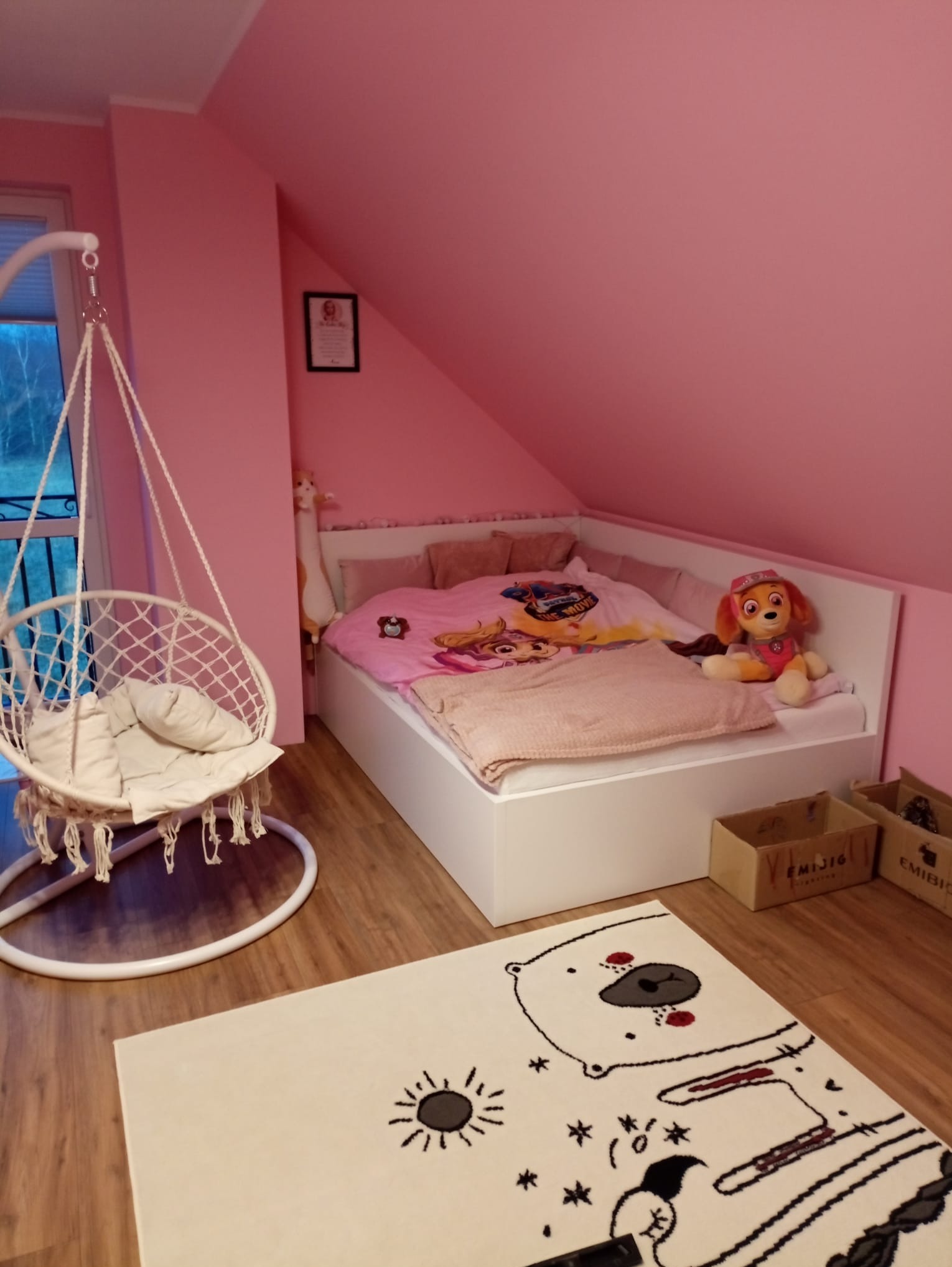 Pokój dziecięcy na poddaszu z różowymi ścianami, białym łóżkiem w zabudowie, wiszącym fotelem hamakowym i dywanem z motywem polarnym.