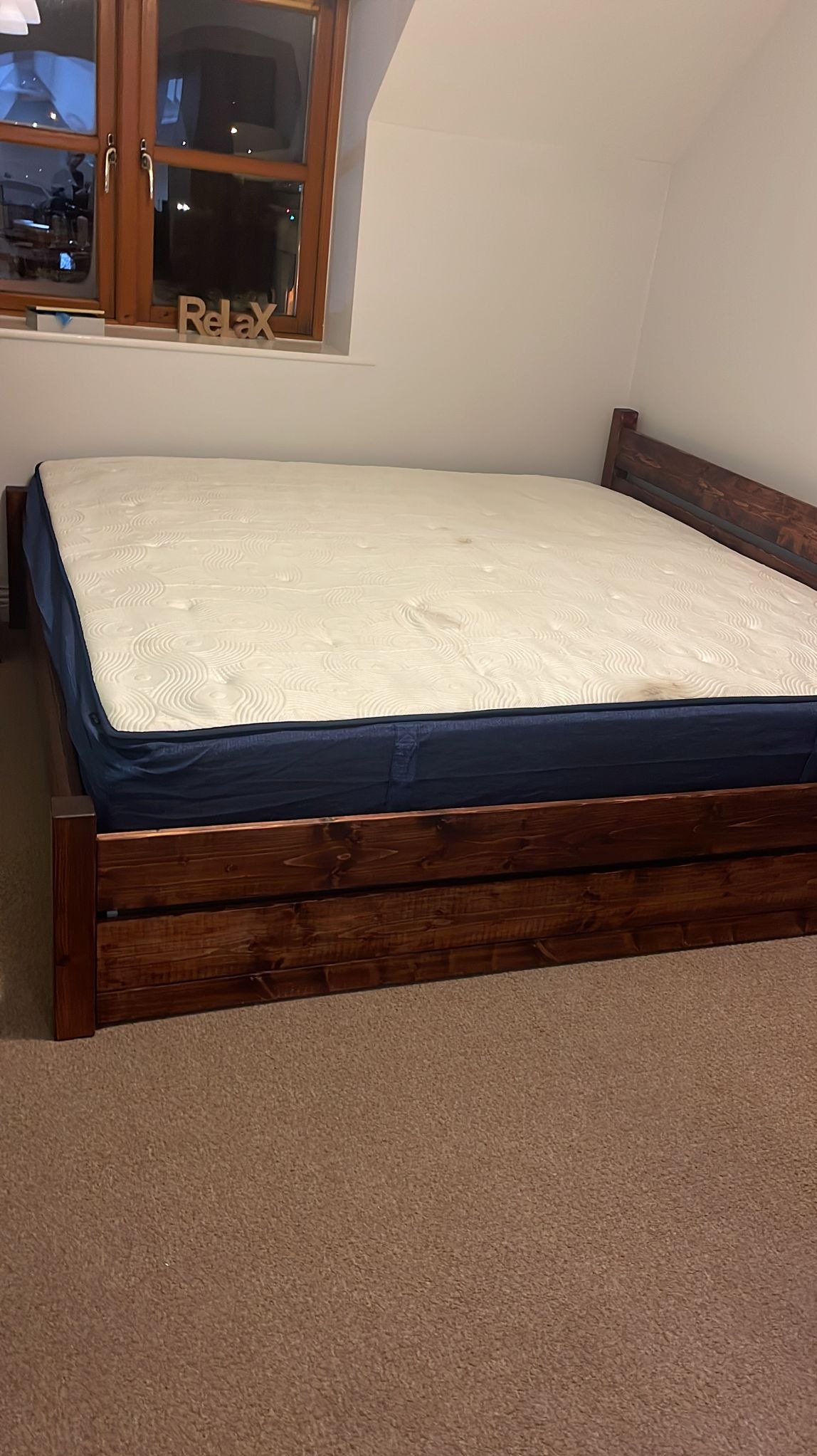 Łóżko z ciemnego drewna z materacem w pokoju z oknem, na parapecie dekoracja 'Relax'