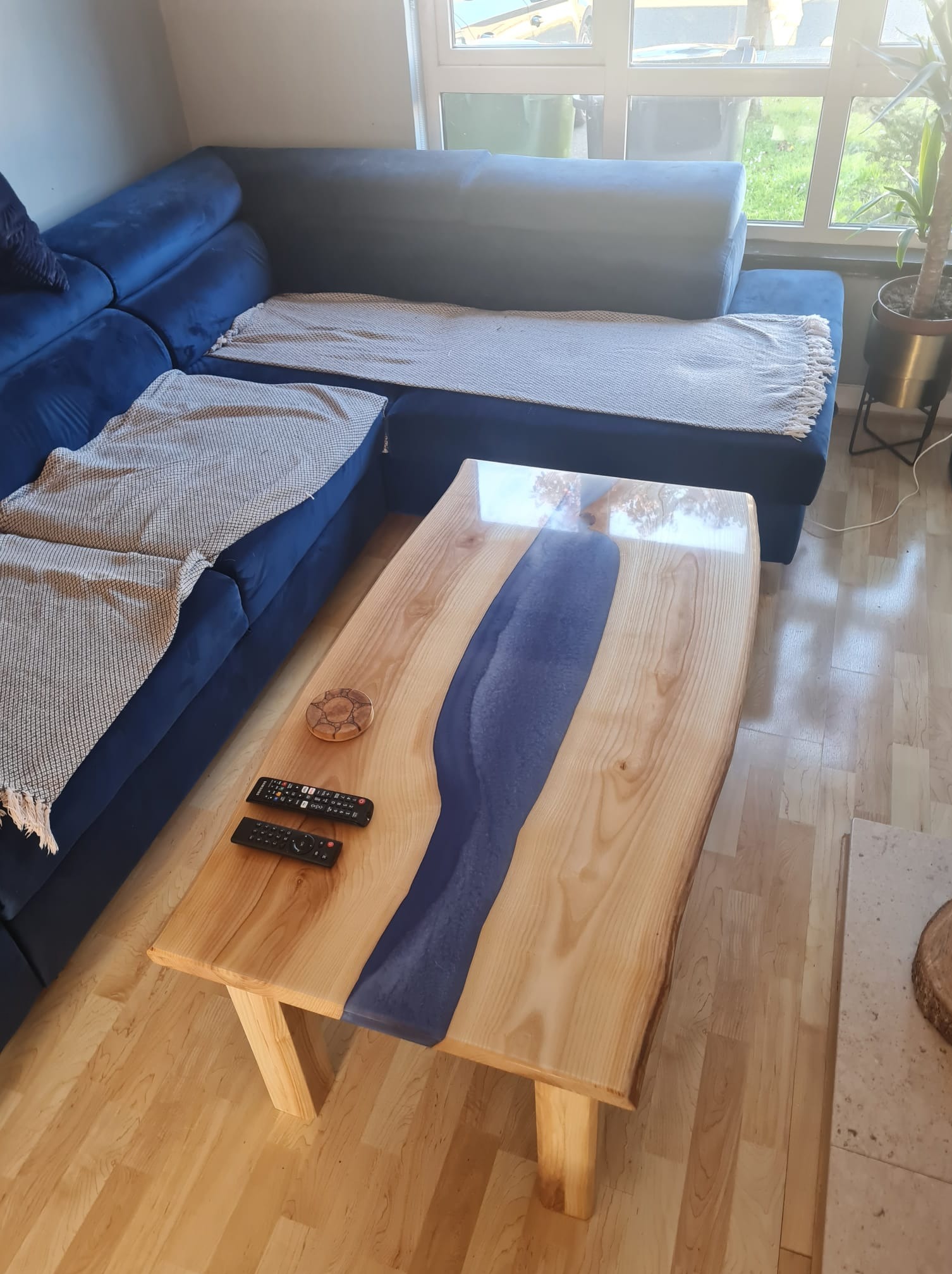 Stolik kawowy z blatem z jasnego drewna z żywicą epoksydową w kolorze granatowym, obok sofy z poduszkami i pilotami.