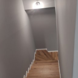 Remontuj Zbigniew Zając - Drewniane schody z białymi listwami przypodłogowymi w szarej klatce schodowej, widok z góry, oświetlone pojedynczym plafonem.