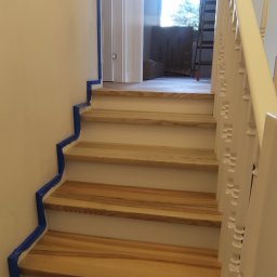 Remontuj Zbigniew Zając - Jasne drewniane schody z białymi podstopnicami i balustradą, zabezpieczone niebieską taśmą malarską przy ścianie, w tle widoczne kartony i drabina
