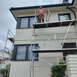 Remontuj Zbigniew Zając - Mężczyzna w koszulce w paski i roboczych spodniach stoi na rusztowaniu przed domem z jasną elewacją, prawdopodobnie podczas prac malarskich.