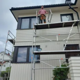 Mężczyzna w koszulce w paski i roboczych spodniach stoi na rusztowaniu przed domem z jasną elewacją, prawdopodobnie podczas prac malarskich.