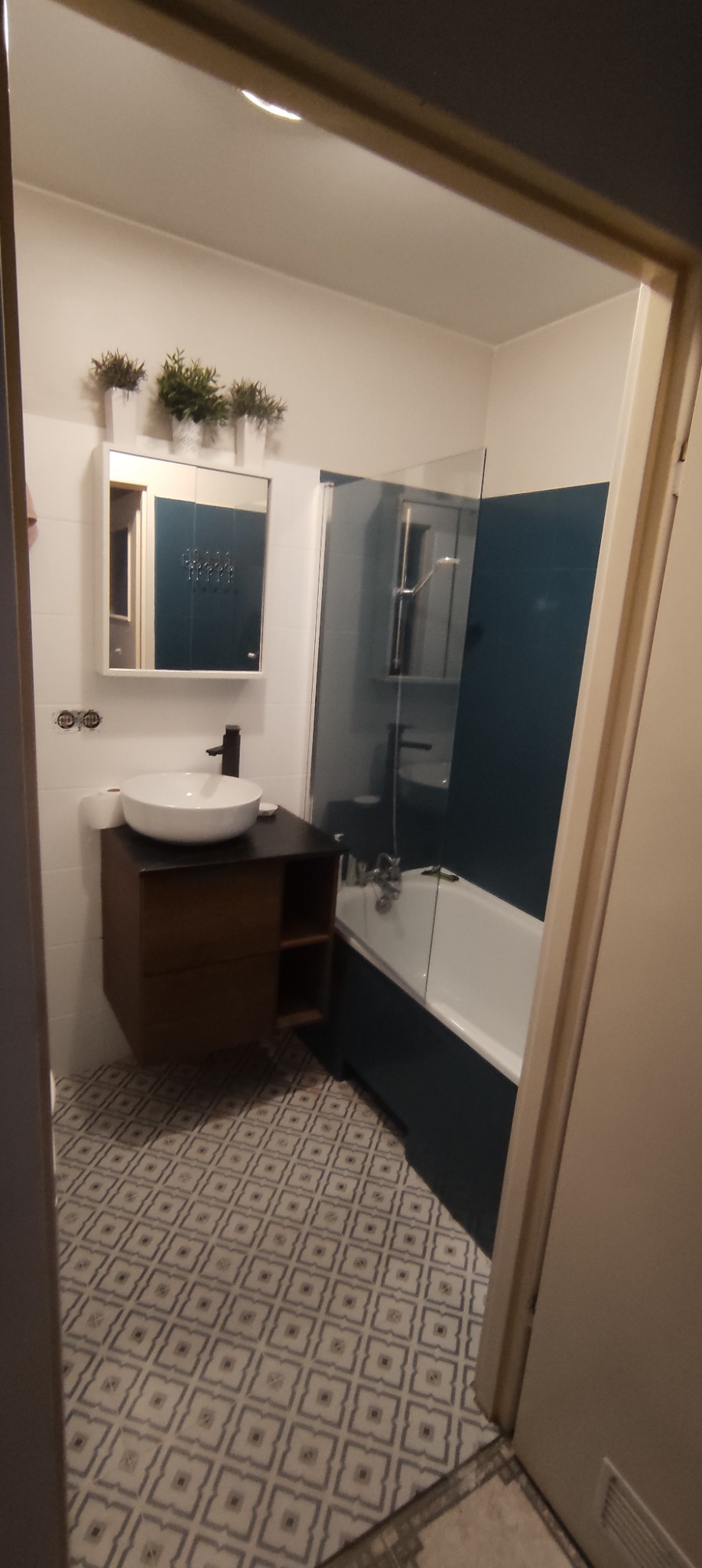 Nowoczesna łazienka z umywalką nablatową na drewnianej szafce, lustrem z szafką, wanną za szklaną ścianką i wzorzystą podłogą, widok zza framugi drzwi.