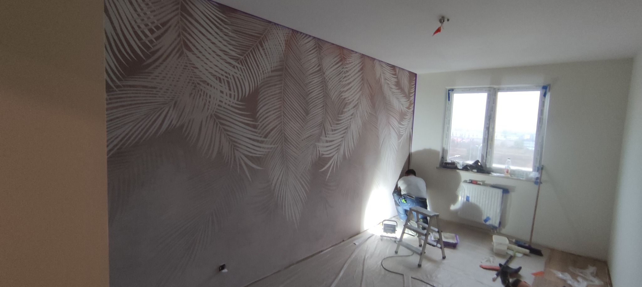 Pracownik na drabinie maluje ścianę w pokoju z wzorem liści palmowych, zabezpieczona podłoga folią malarską, widoczne narzędzia malarskie i oświetlenie robocze.