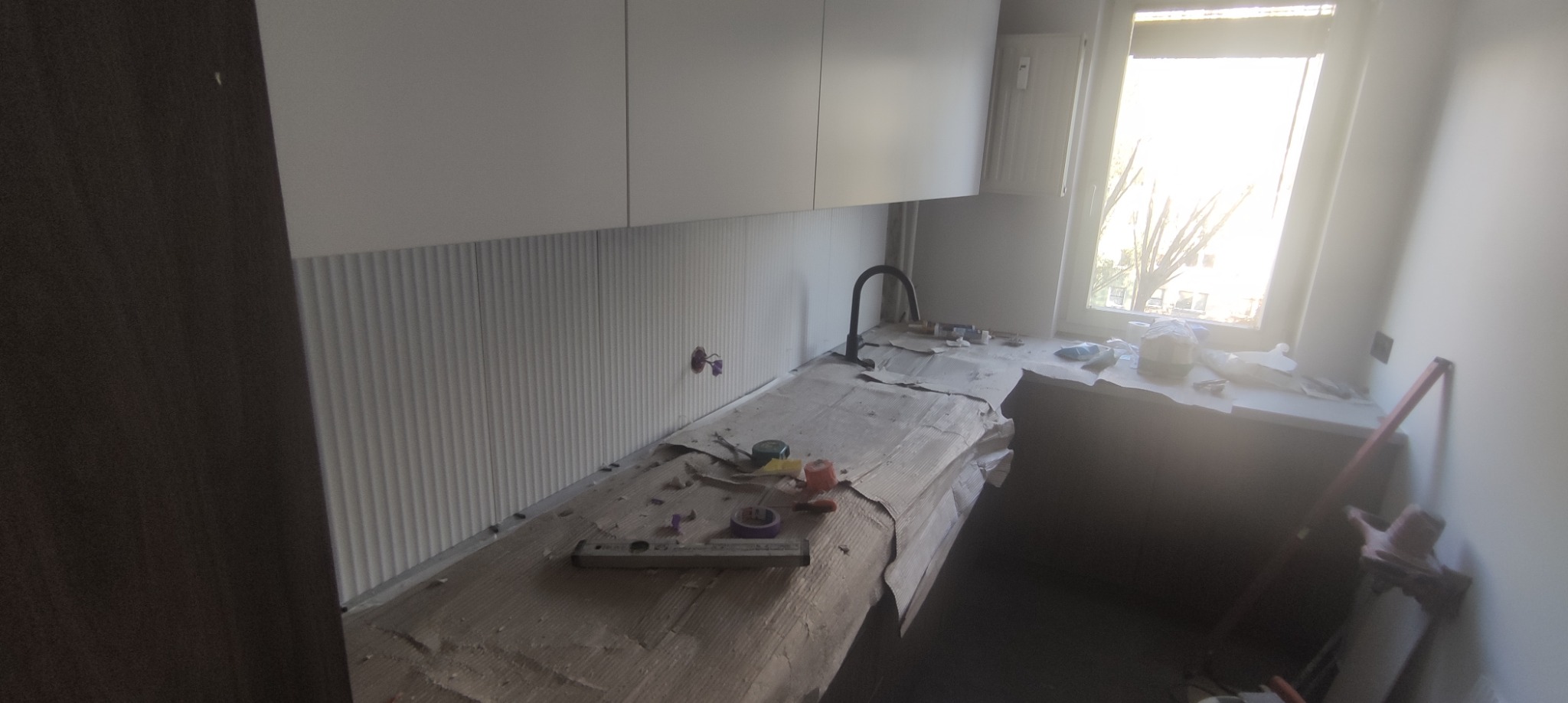 Montaż paneli ściennych z pionowymi żłobieniami w kuchni, widoczne narzędzia i zakryty blat, okno z widokiem na drzewo