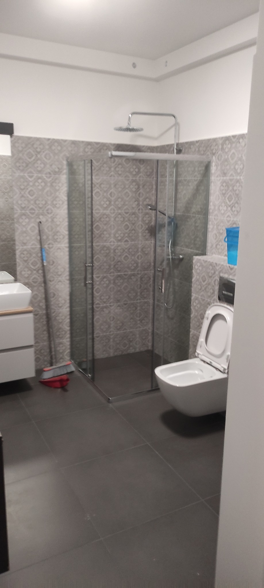 Wnętrze łazienki z kabiną prysznicową typu walk-in, umywalką na szafce i wiszącą toaletą z otwartą klapą, szare płytki na ścianach i podłodze, widoczna szczotka do sprzątania.