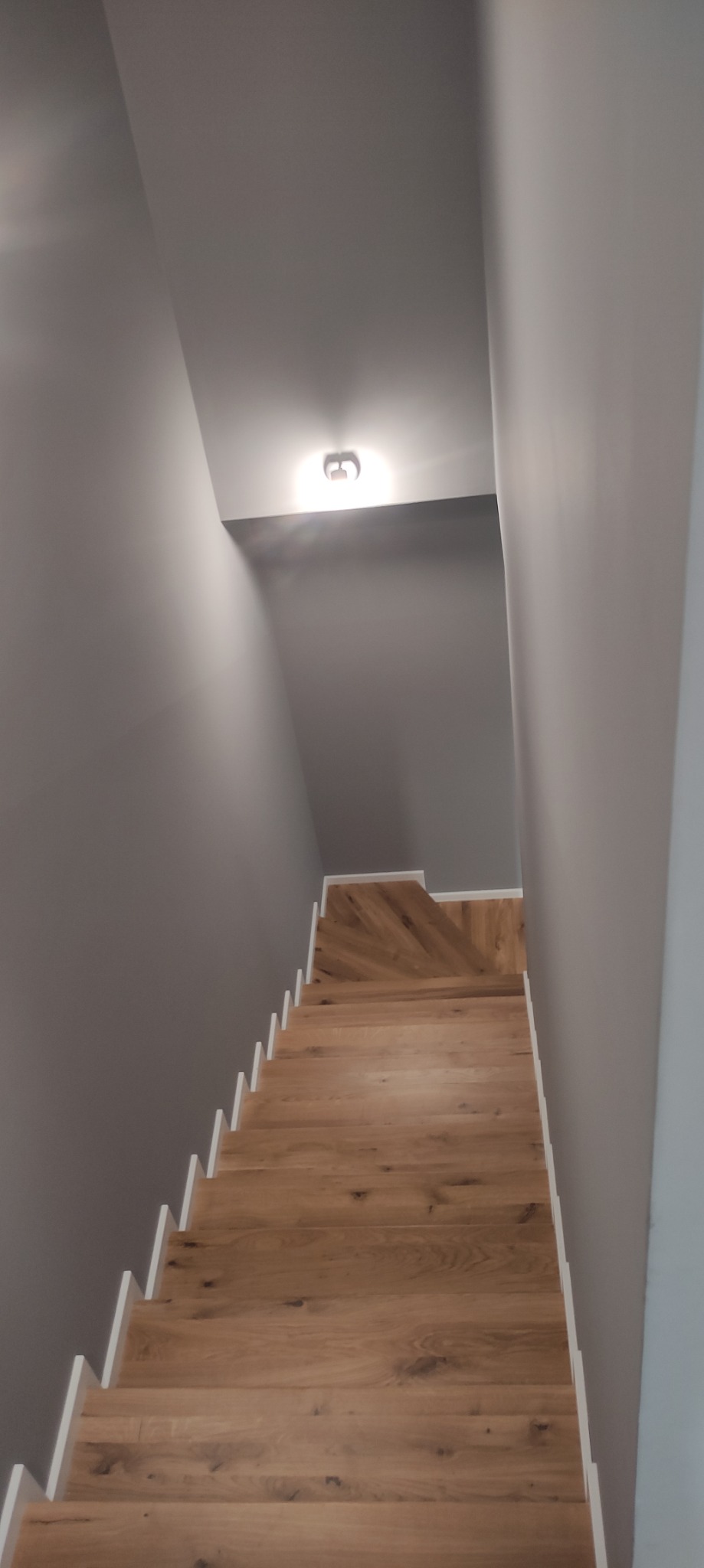 Drewniane schody z białymi listwami przypodłogowymi w szarej klatce schodowej, widok z góry, oświetlone pojedynczym plafonem.