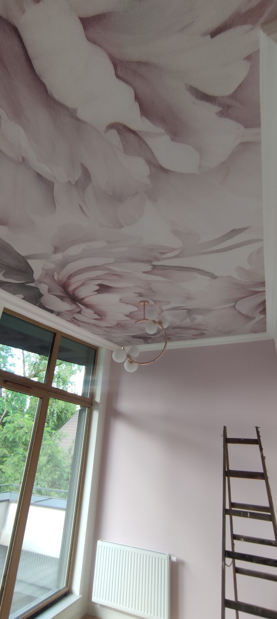 Różowe kwiaty na suficie w pokoju z oknem, grzejnikiem i drabiną, oświetlone dekoracyjną lampą