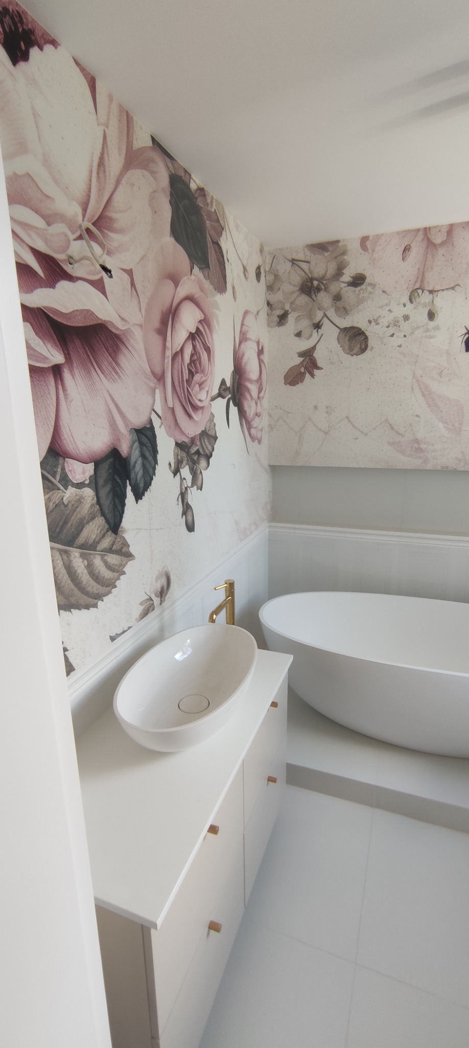 Biała owalna umywalka nablatowa z złotą baterią, umieszczona na białej szafce z minimalistycznymi uchwytami, w tle wolnostojąca wanna i ściany z motywem kwiatowym w odcieniach różu i szarości.
