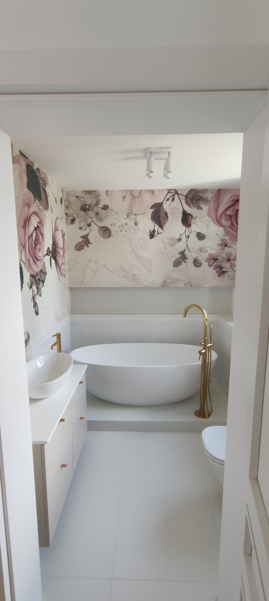 Biała łazienka z owalną wanną, umywalką nablatową na szafce i tapetą w kwiaty róży, złota armatura, minimalistyczne oświetlenie sufitowe.