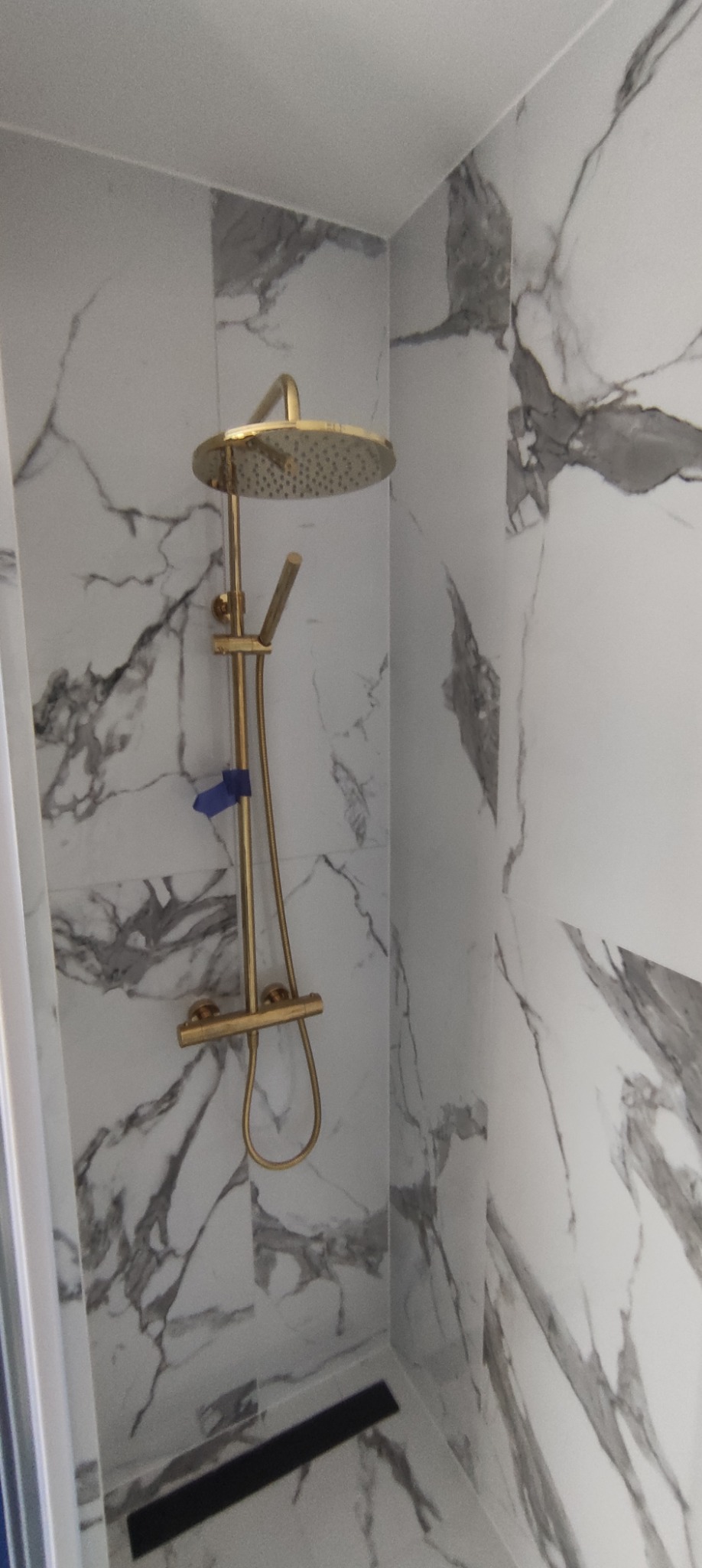 Złota bateria prysznicowa z deszczownicą na tle marmurowych płytek w kabinie prysznicowej w Józefowie, z widocznym odpływem liniowym.