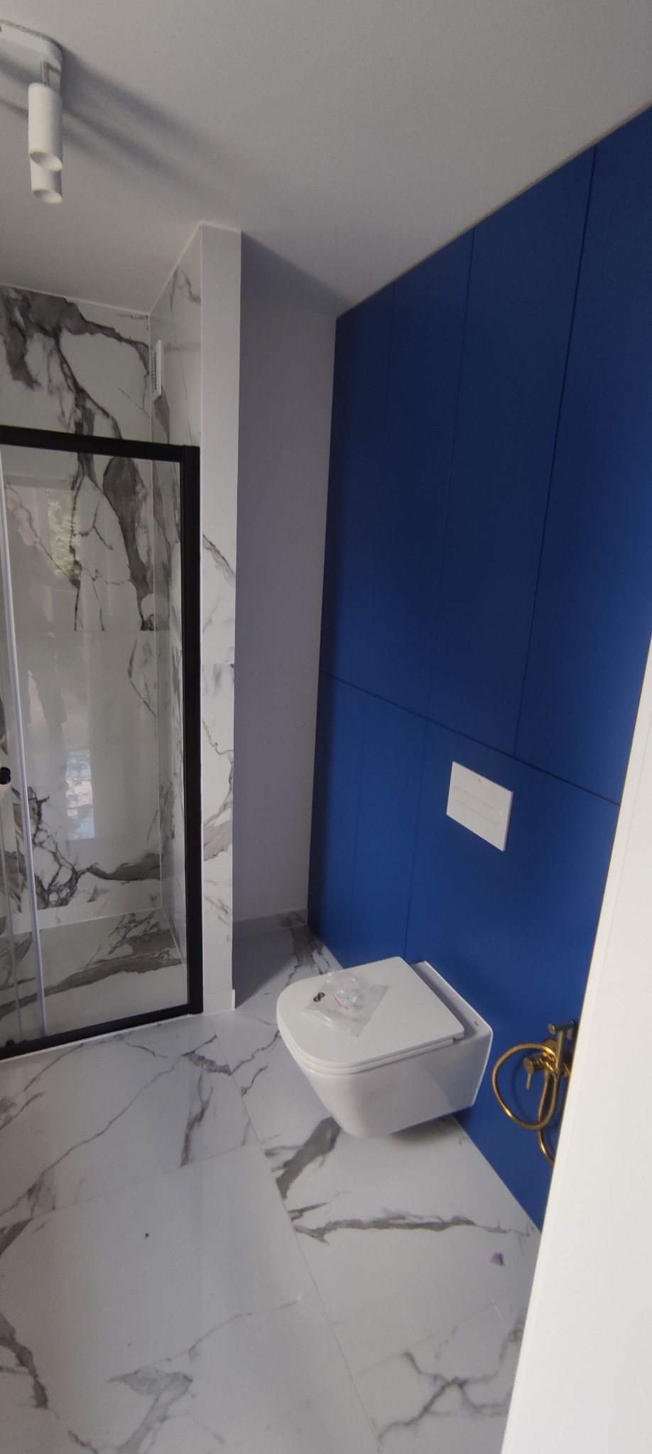 Nowoczesna łazienka z kabiną prysznicową w czarnej ramie, podwieszaną toaletą z opakowaniem montażowym na desce oraz zabudową w kolorze ciemnego błękitu.