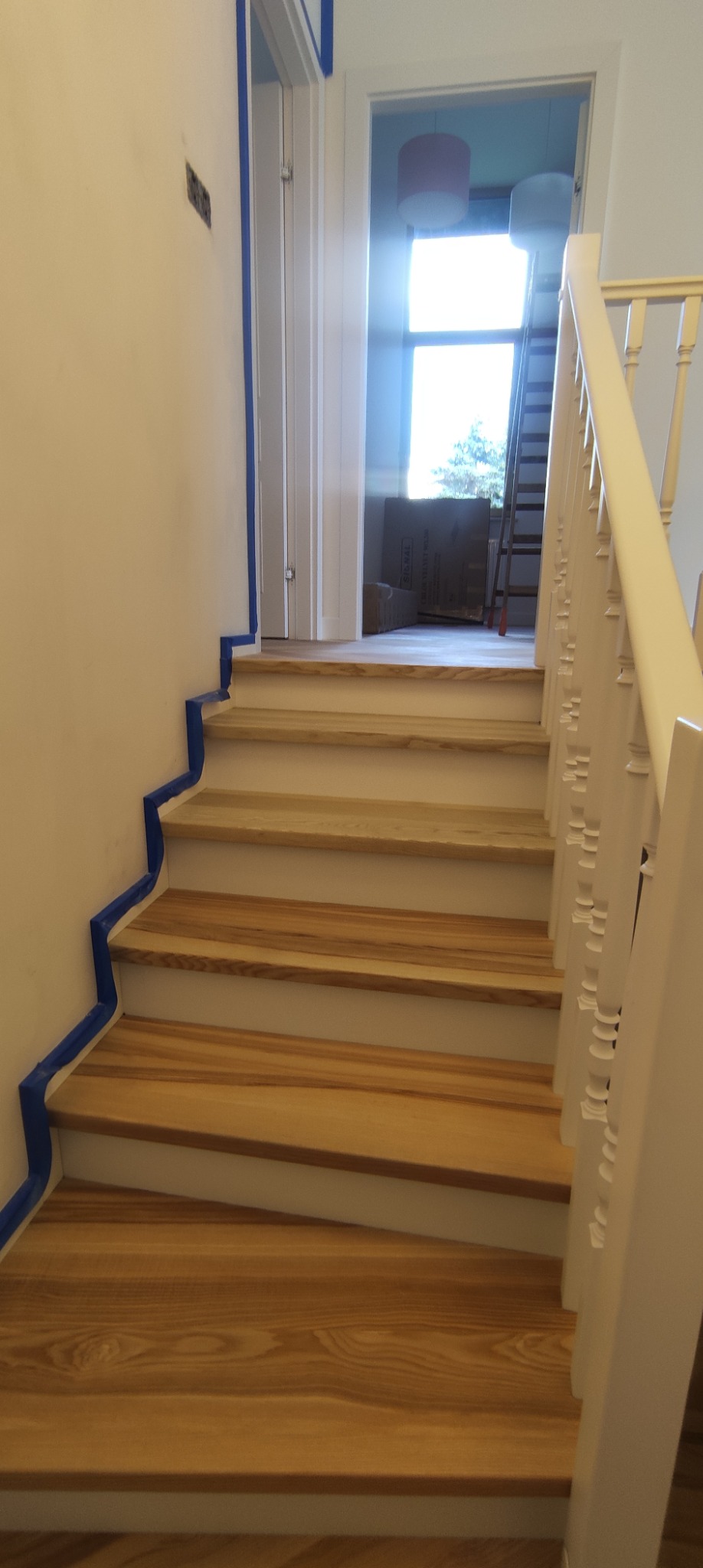 Jasne drewniane schody z białymi podstopnicami i balustradą, zabezpieczone niebieską taśmą malarską przy ścianie, w tle widoczne kartony i drabina