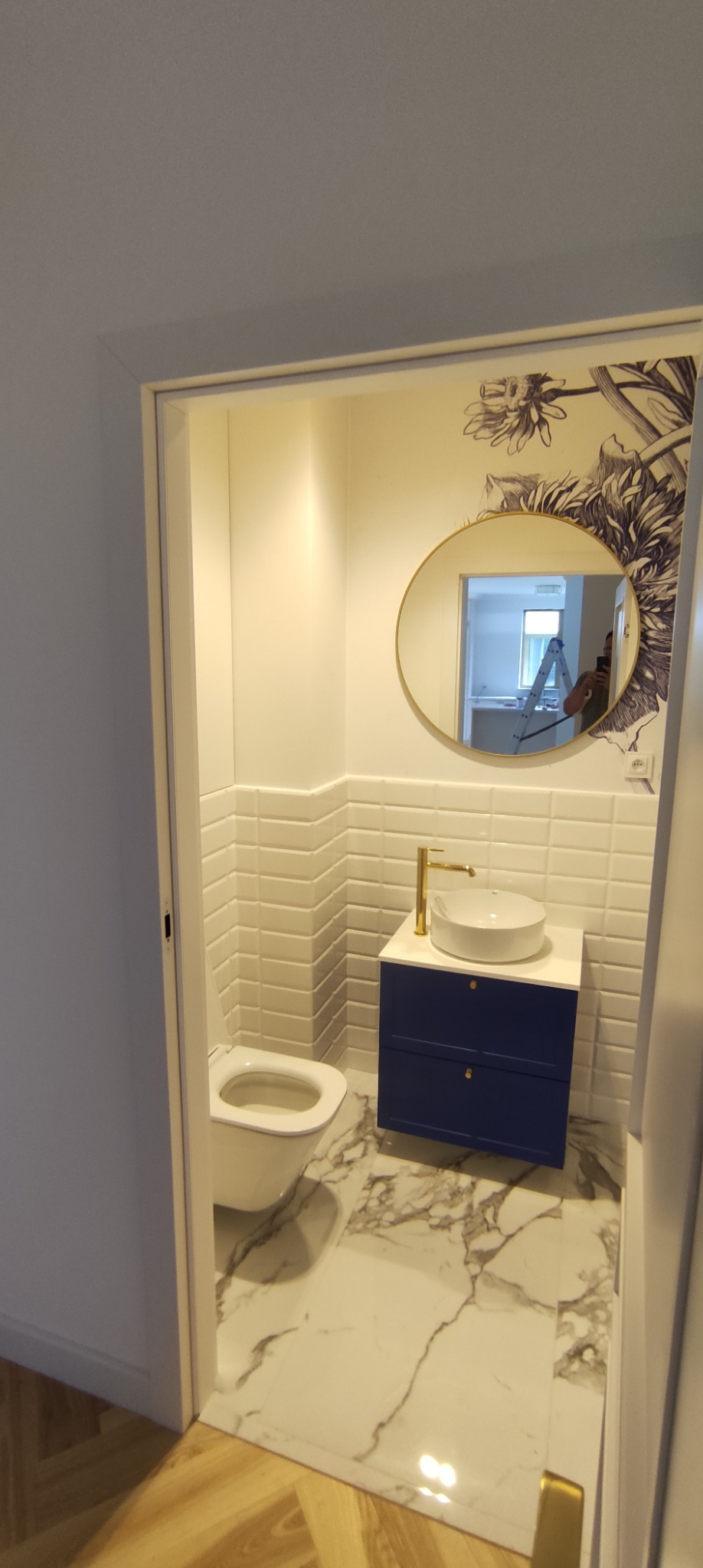 Nowoczesna łazienka z białą ceramiką, granatową szafką pod umywalką z złotą armaturą i marmurową podłogą, widok przez otwarte drzwi, na ścianie dekoracyjny motyw roślinny oraz okrągłe lustro.