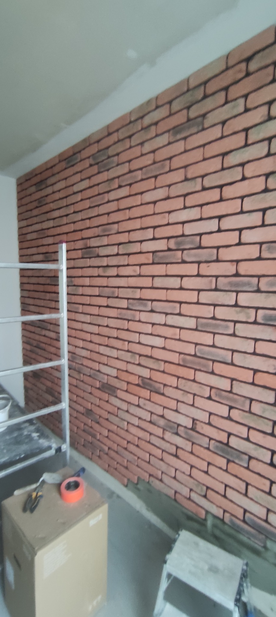 Ściana wyłożona dekoracyjną cegłą, widoczny fragment rusztowania i narzędzia na kartonie, etap prac wykończeniowych.