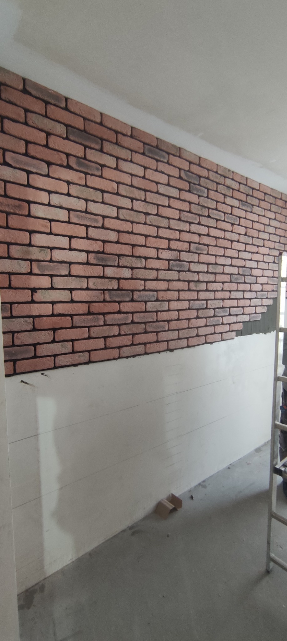 Montaż dekoracyjnej ściany z cegieł na częściowo wykończonej ścianie, widoczny fragment drabiny.
