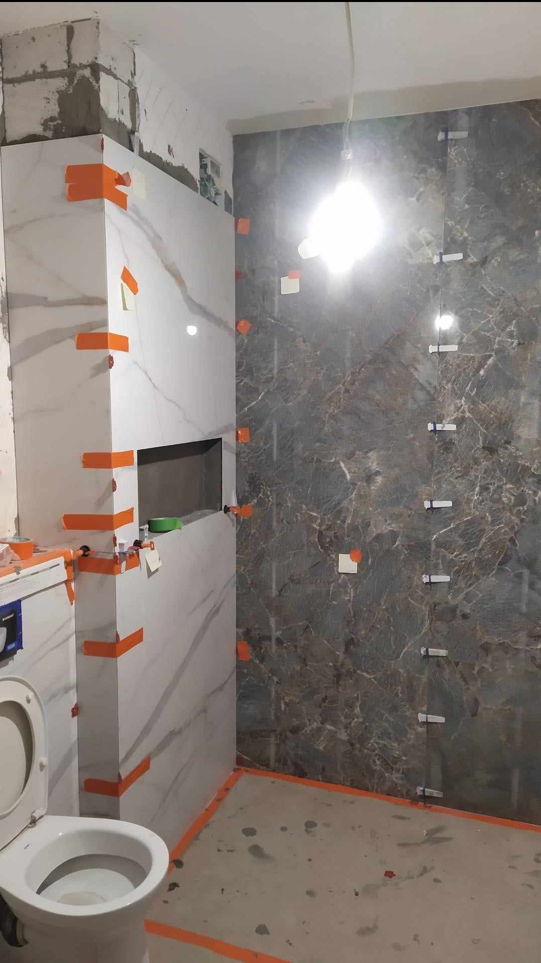 Montaż płytek imitujących marmur w łazience, widoczne pomarańczowe paski taśmy i plastikowe krzyżyki dystansowe, surowy stan podłogi.
