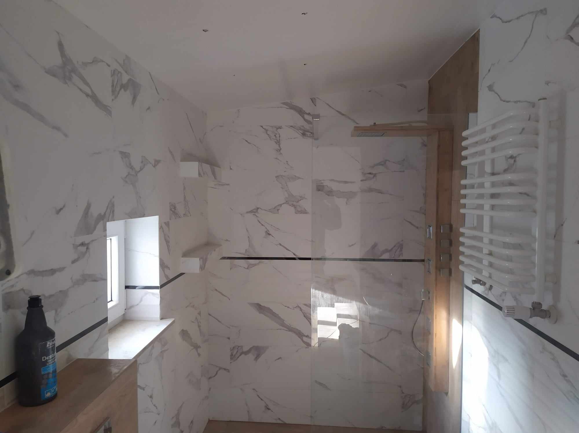 Wykończona łazienka z białymi płytkami imitującymi marmur, kabina prysznicowa ze szklaną ścianką, grzejnik drabinkowy i butelka środka czyszczącego na blacie pod oknem.