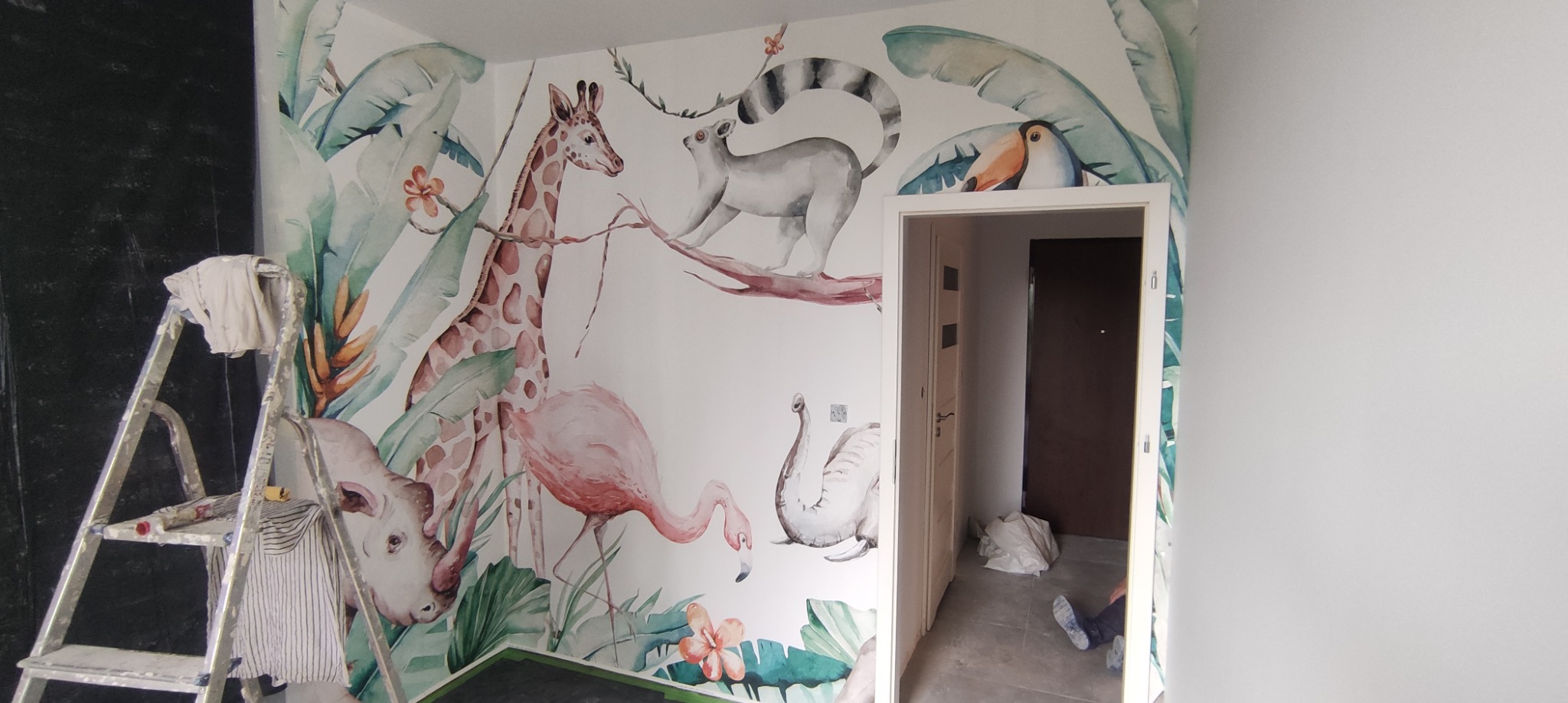 Ściana pokoju dziecięcego ozdobiona tapetą z motywem zwierząt tropikalnych, takich jak żyrafa, lemur, tukan, flaming i nosorożec, z widoczną drabiną malarską i częściowo pomalowaną czarną ścianą...