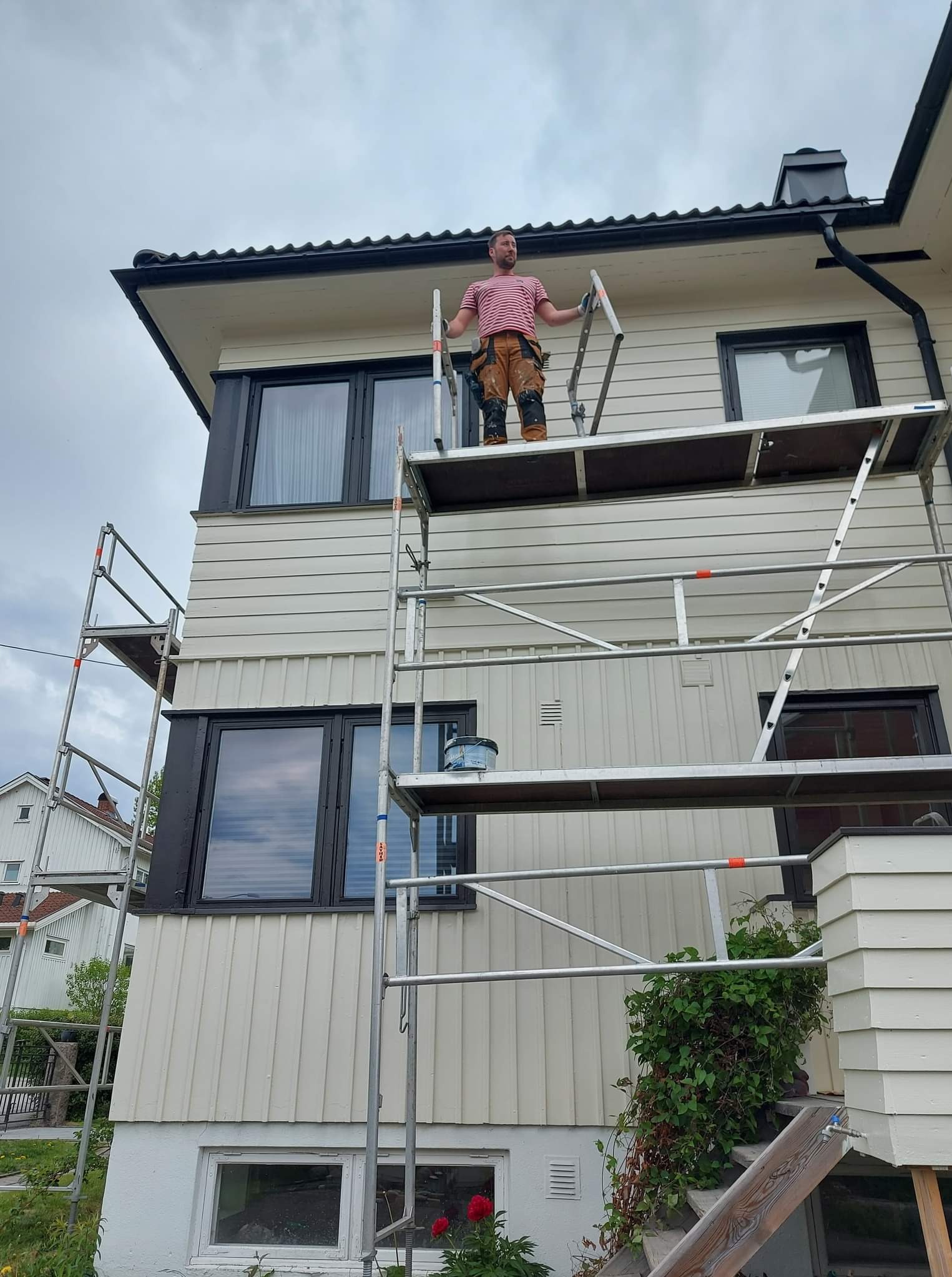 Mężczyzna w koszulce w paski i roboczych spodniach stoi na rusztowaniu przed domem z jasną elewacją, prawdopodobnie podczas prac malarskich.