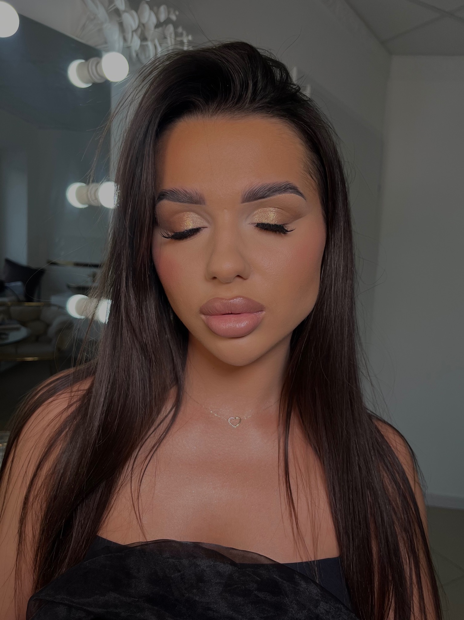 Portret kobiety z zamkniętymi oczami, prezentującej złoty odcień cieni do powiek, wykonturowane policzki i błyszczące usta, z delikatnym naszyjnikiem w kształcie serca, na tle lustra z oświetleniem.